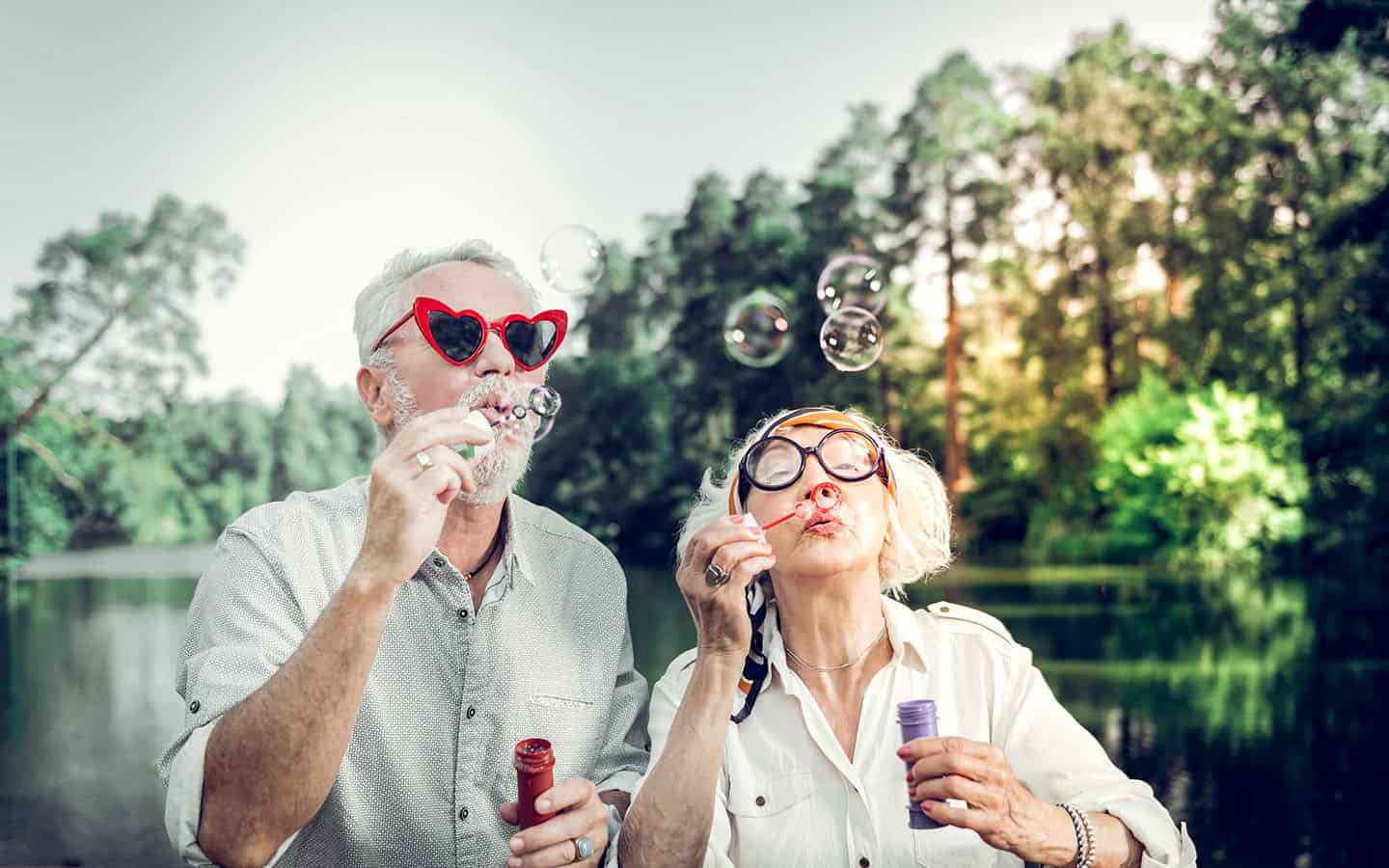 blog_hundertjaehrige A cheerful senior couple outdoors by a lake blowing soap bubbles, with the man wearing heart-shaped red sunglasses and the woman wearing large round glasses.