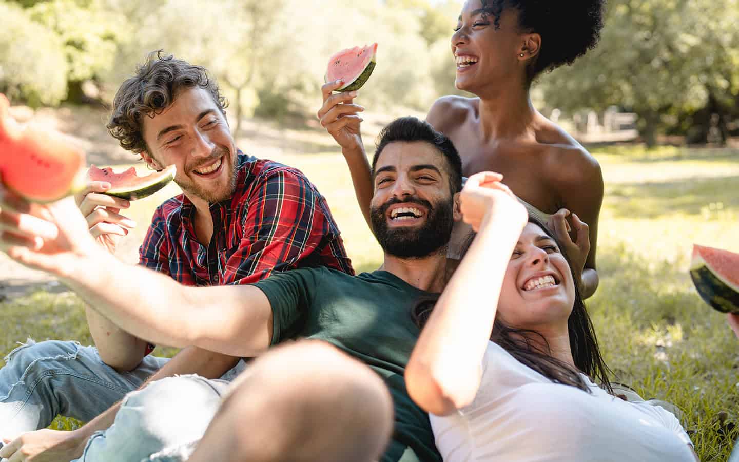 blog_lebensstil A group of diverse friends sitting together on the grass in a sunny park, laughing and enjoying fresh watermelon slices.