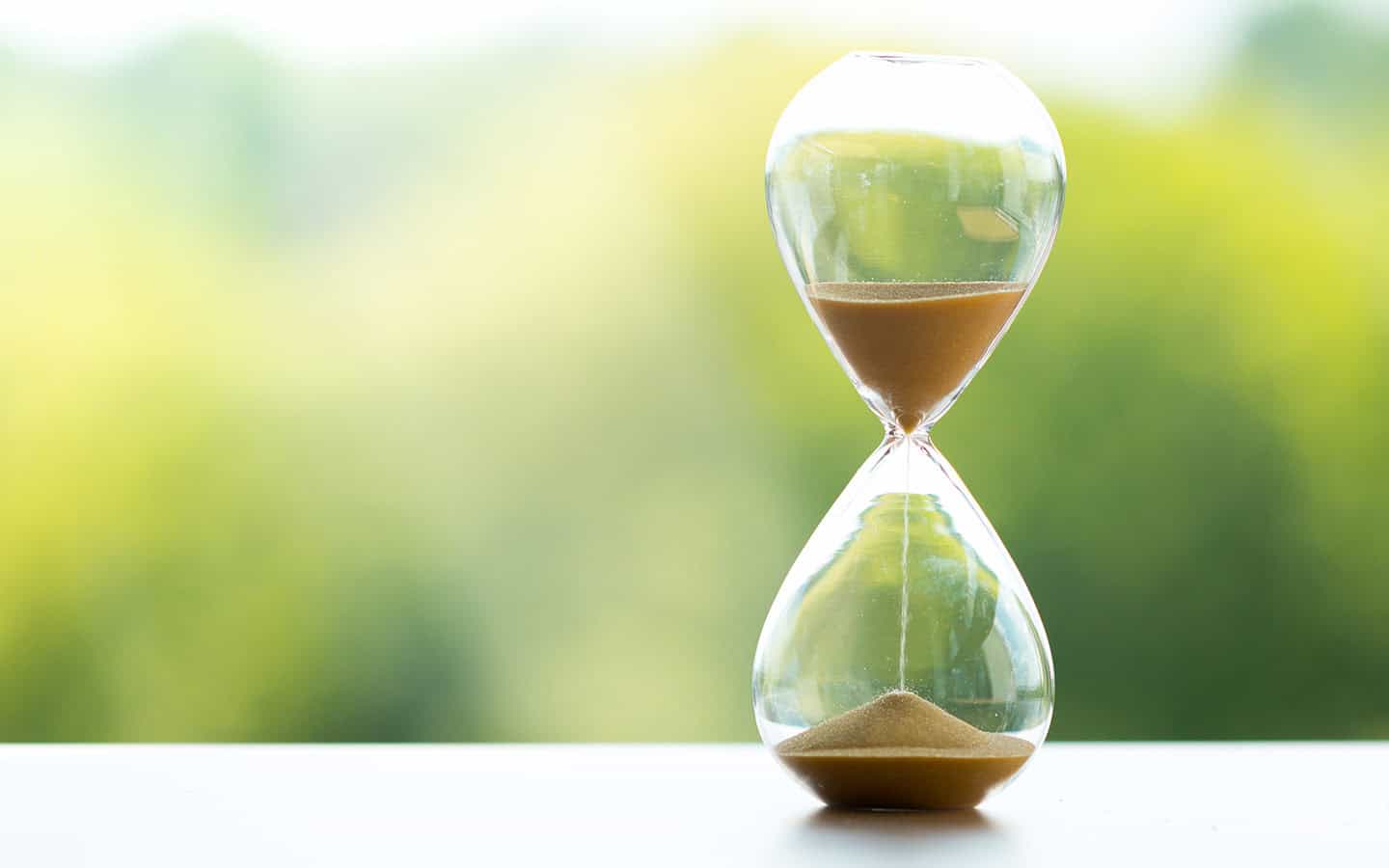 blog_maximale_lebenserwartung A glass hourglass filled with fine golden sand trickling through its narrow neck, resting on a white surface against a soft-focus green natural background.